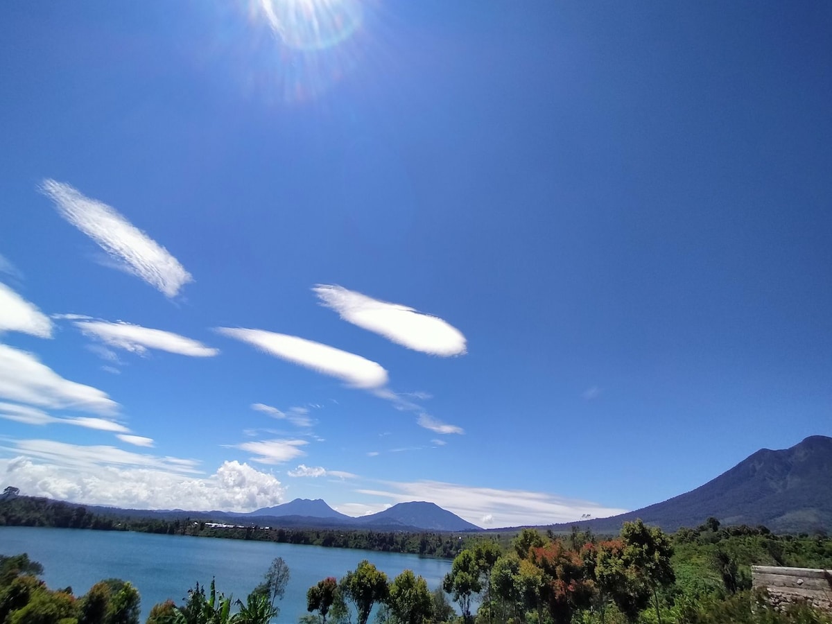 A panoramic view showcases a clear blue sky adorned with wispy clouds above a serene lake. Three mountains are visible in the background, framed by lush greenery along the water's edge, creating a peaceful natural landscape.