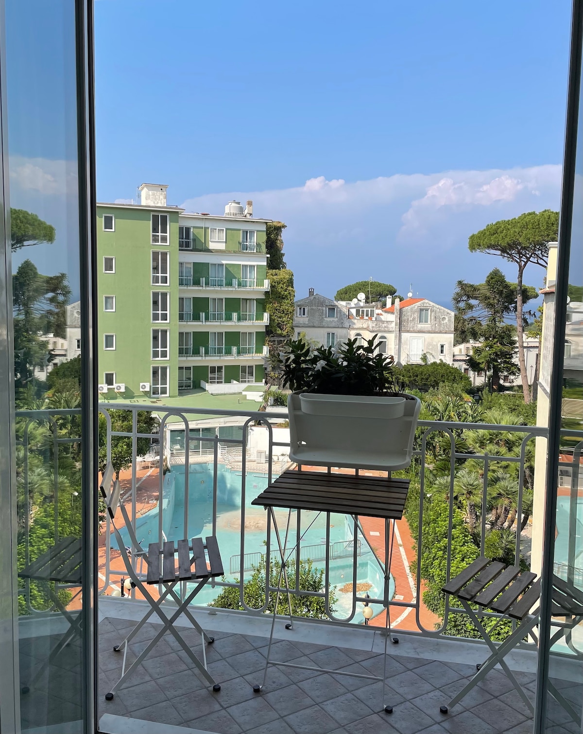 A balcony view captures a quiet pool area surrounded by lush greenery, with a modern green building visible in the background. Two small folding chairs are positioned on the balcony, providing a space to relax and enjoy the scenic surroundings.