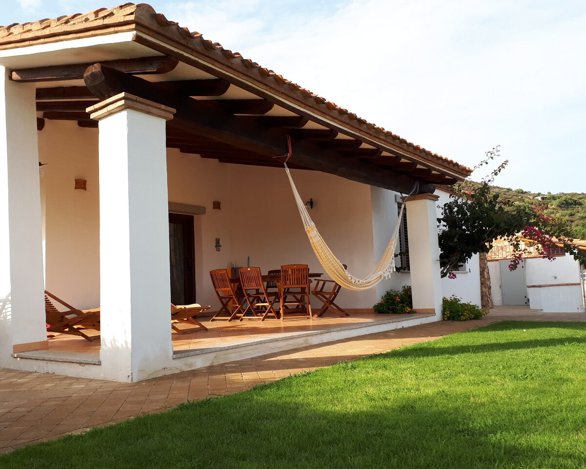 A spacious covered terrace features wooden dining furniture and a hammock, surrounded by a well-maintained lawn. The structure’s traditional roof provides shade, while the serene landscape in the background enhances the outdoor relaxation space.