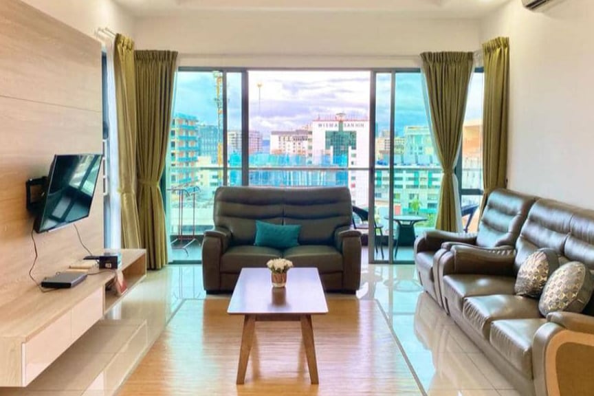 The living room features large windows offering city and sea views. A flat-screen TV is mounted on the wall, with a wooden coffee table centrally placed. Two comfortable leather sofas are arranged to foster conversation, complemented by curtains that frame the scenic backdrop.