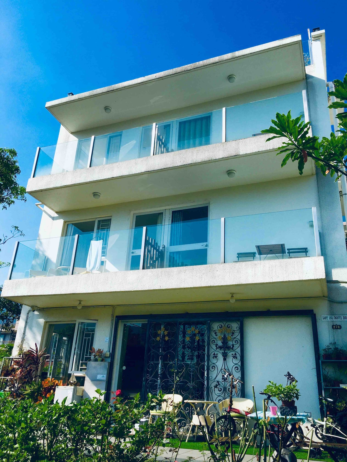 The three-story villa features a modern façade with large glass balconies and an ornate entrance gate. Greenery surrounds the base of the building, enhancing the outdoor space. Clear blue skies provide a bright backdrop, highlighting the architectural details.