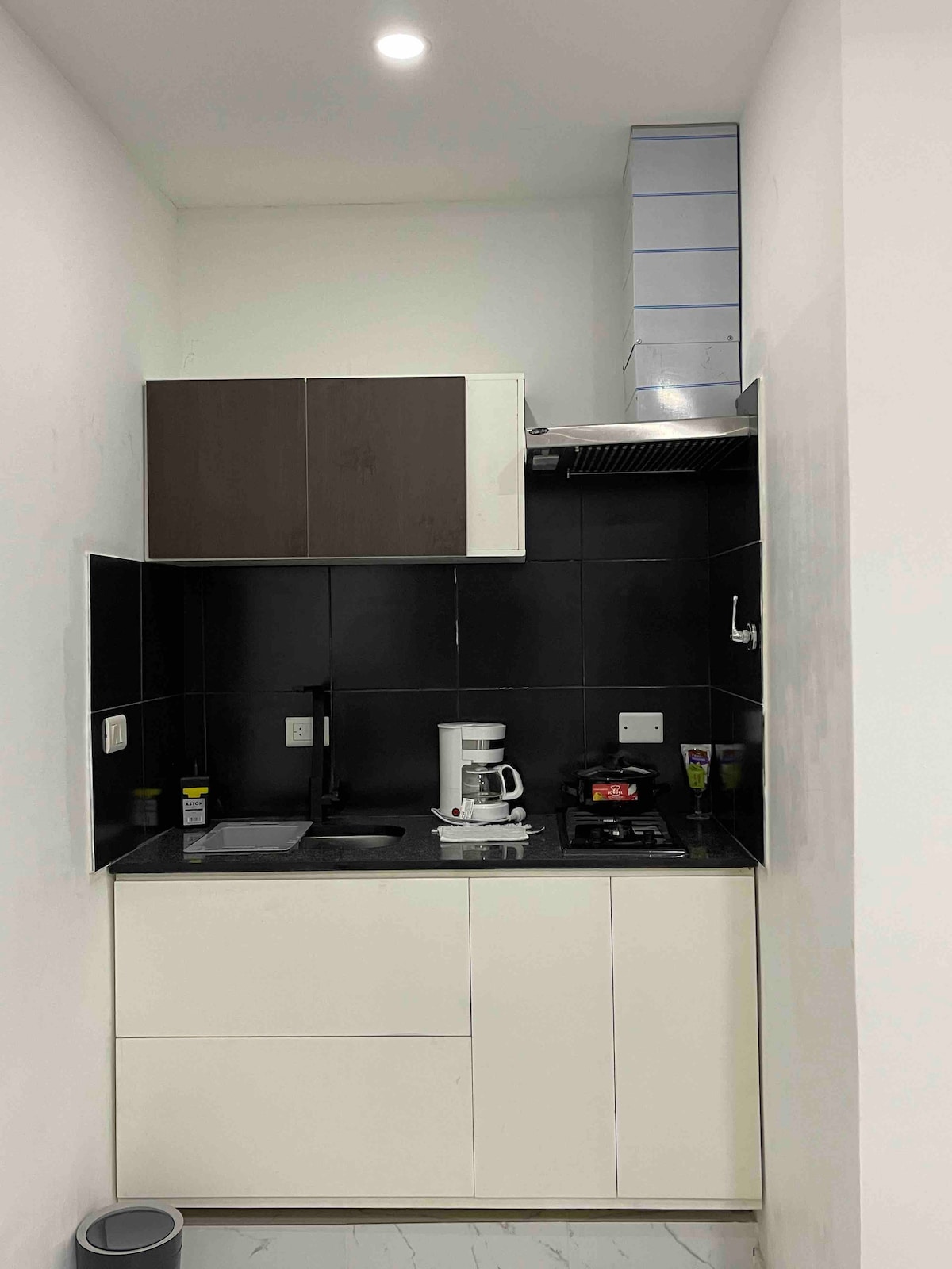 A compact kitchen is presented with modern cabinetry in contrasting shades of black and white. A coffee maker and stovetop are visible, along with essential cooking utensils. The countertops are clear, enhancing the kitchen's functional design. Bright lighting illuminates the space, highlighting the sleek surfaces.