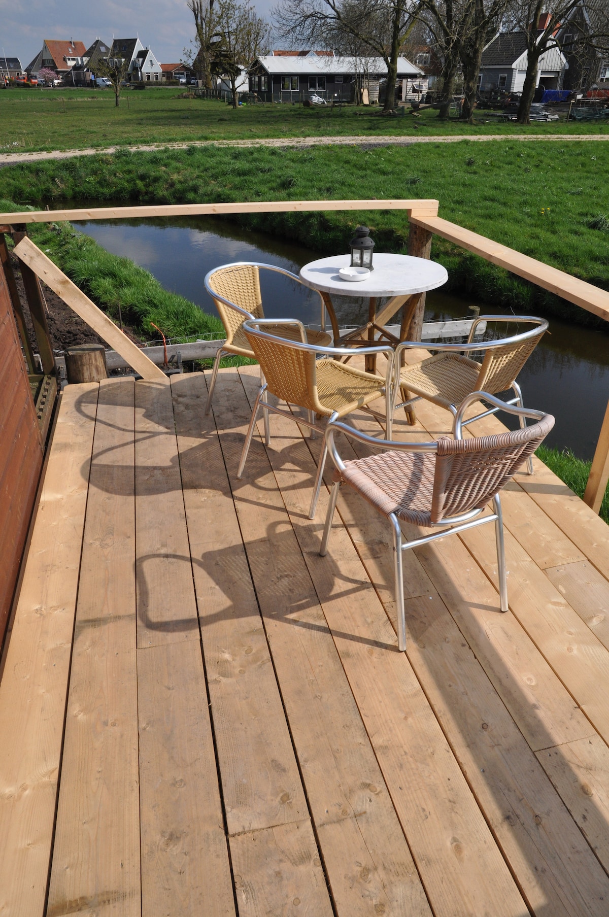 An outdoor terrace is presented, featuring a round tabletop accompanied by four chairs. The wooden deck offers a natural finish, and a scenic view of a grassy area and a small waterway is visible nearby.