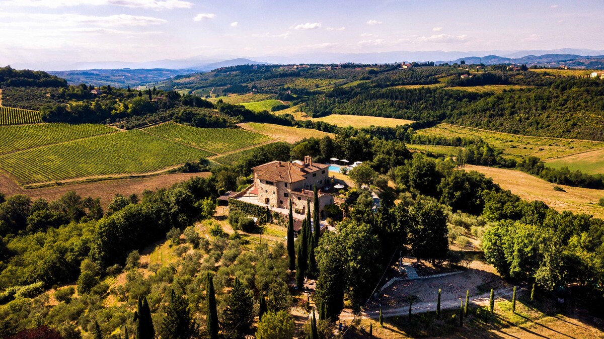 An aerial view captures the cottage surrounded by lush green hills and vineyards. The expansive landscape features a patchwork of fields and trees, emphasizing the serene countryside setting. The structure is framed by tall cypress trees, enhancing the tranquil atmosphere of the location.