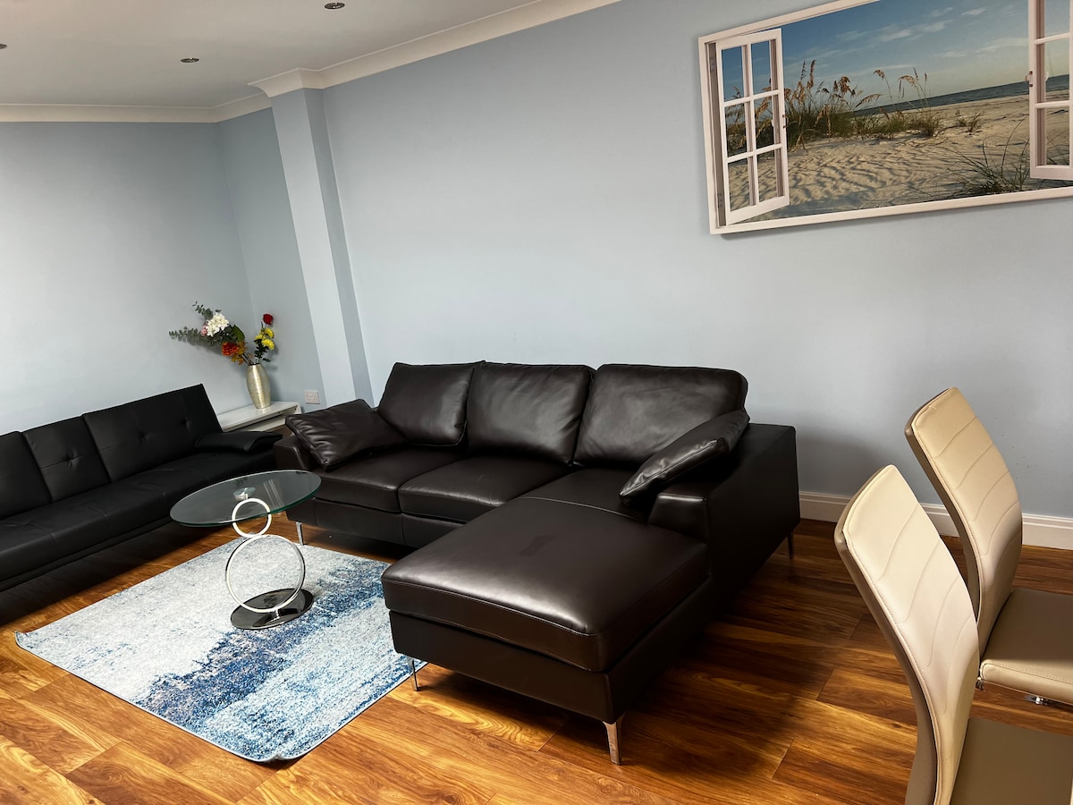 A spacious lounge area features a dark leather sectional sofa positioned alongside a glass coffee table. A light blue area rug contrasts with the warm wooden floors. An artwork depicting a beach scene is displayed on the wall above the sofa.
