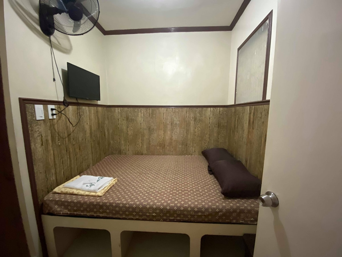 A compact sleeping area features a spacious bed with two pillows and a folded towel placed at the foot. The room includes a wall-mounted television and a ceiling fan, with wooden paneling partially surrounding the bed, creating a simple yet functional design.