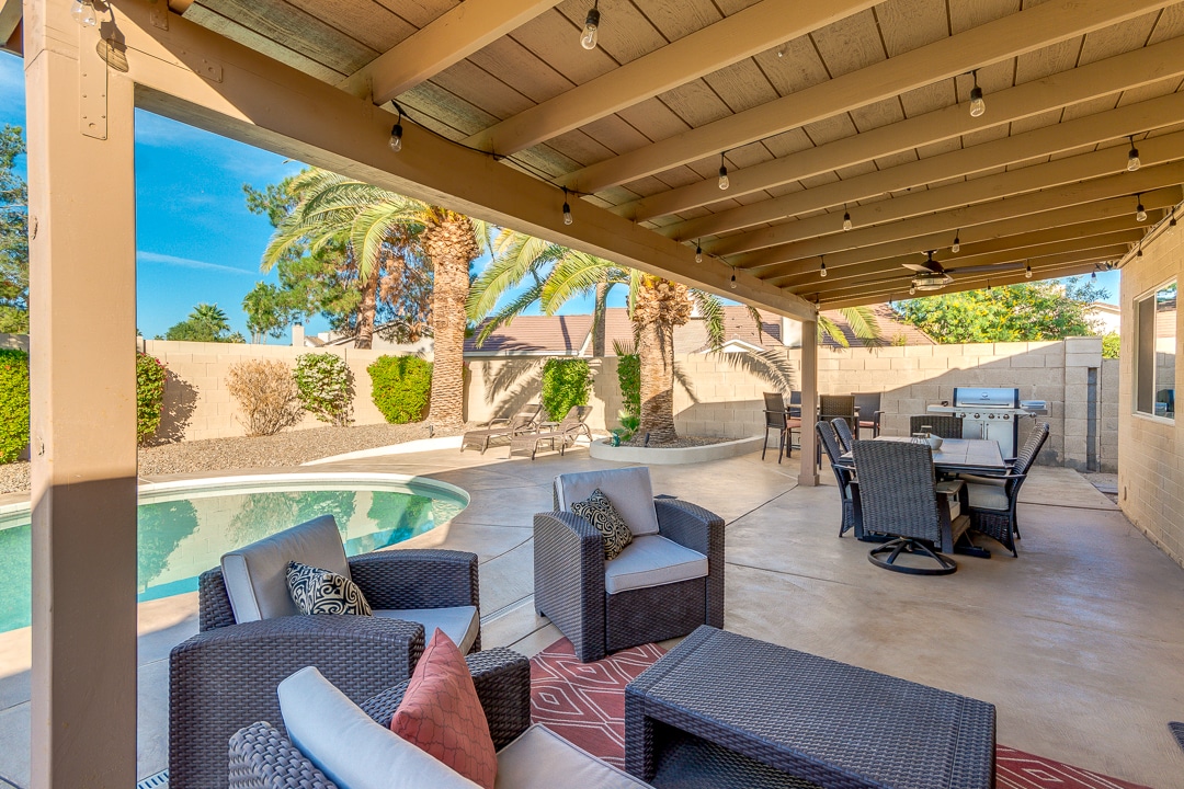 An outdoor lounge area features comfortable seating with soft cushions, arranged under a shaded patio. An adjacent dining table is visible, while the private pool glistens in the sunlight. Lush greenery and palm trees surround the space, enhancing the relaxed environment.