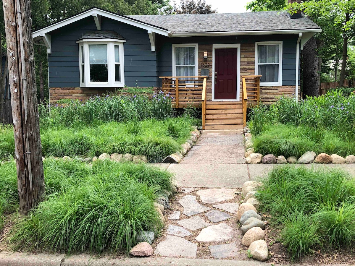 A charming home exterior is framed by lush green grass and flower beds. A stone pathway leads to a wooden front porch with railings and a red front door, inviting guests to enter. Tall trees provide shade in this serene residential setting.