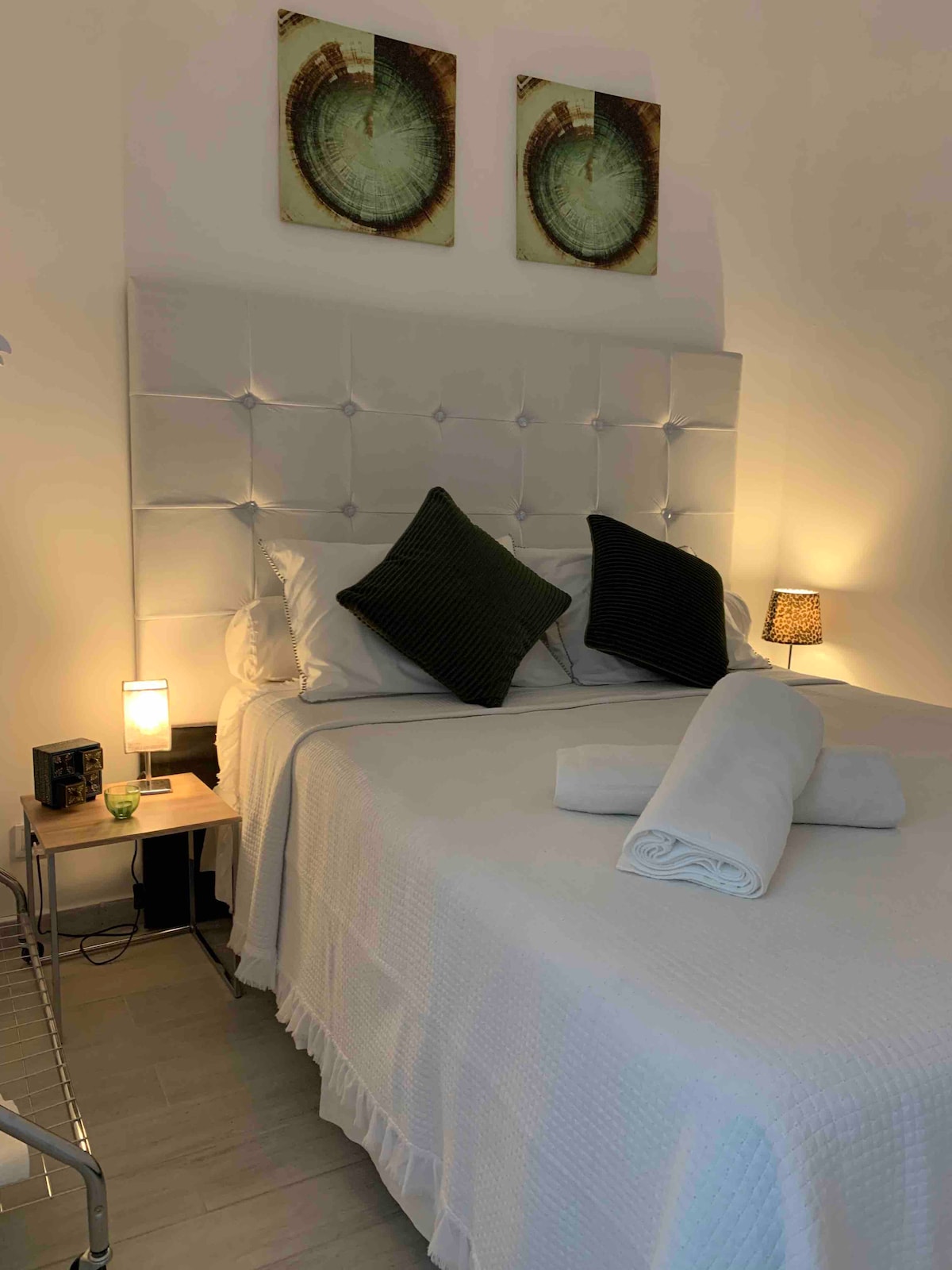 A cozy sleeping area features a light-colored bed adorned with a tufted headboard. Two dark green decorative pillows rest atop the bedspread. Warm lighting is provided by a bedside lamp, and rolled towels are neatly placed at the foot of the bed.