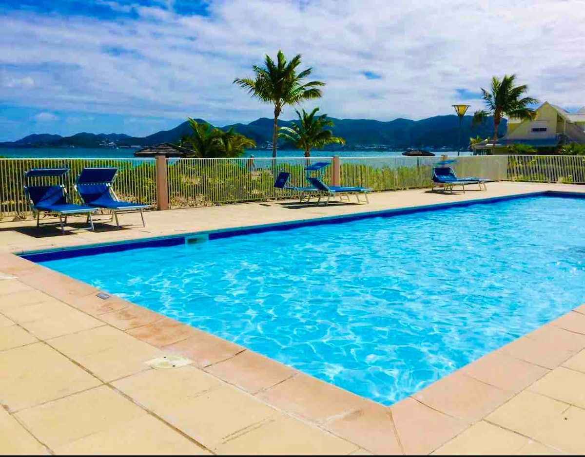 A clear blue pool is surrounded by a tiled deck, with several lounge chairs positioned nearby. Lush palm trees and distant mountains provide a scenic backdrop against a partly cloudy sky. The pool area invites relaxation and enjoyment of the view.
