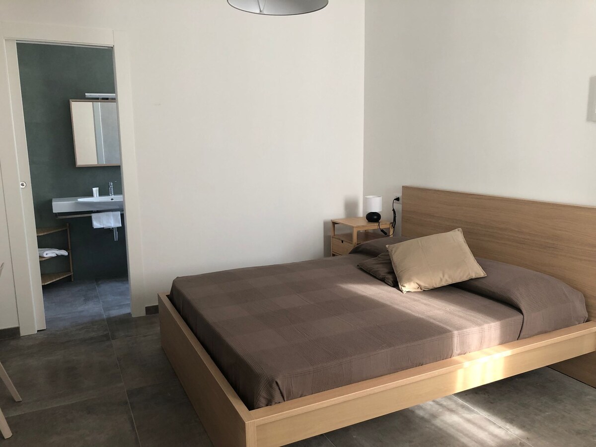 A sleeping area features a wooden bed with a neutral-colored blanket and a decorative pillow. Soft, natural light enters through a window, illuminating the minimalist design. A wooden bedside table holds a lamp, and a bathroom area is visible in the background.