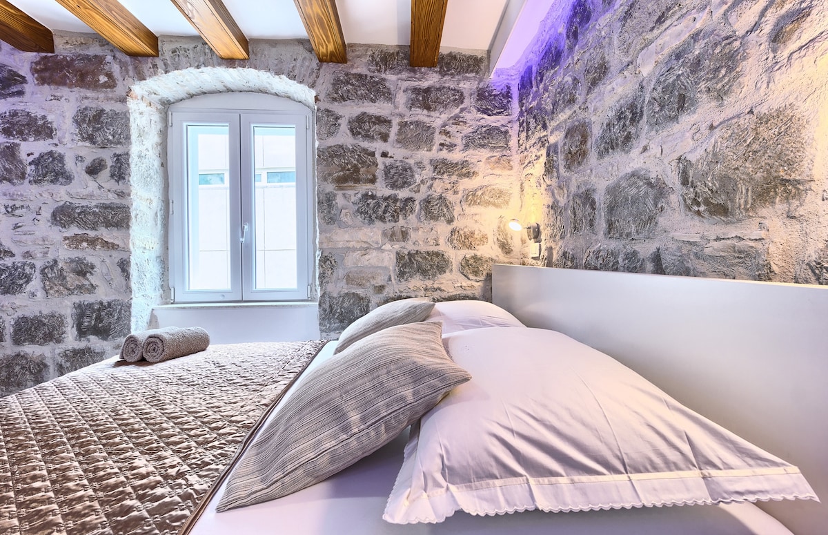 A cozy sleeping area showcases a modern bed with soft linens and two decorative pillows. The stone wall provides a textured backdrop, while light accents illuminate the space. A window is partially visible, allowing natural light to enter, and towels are neatly arranged nearby.