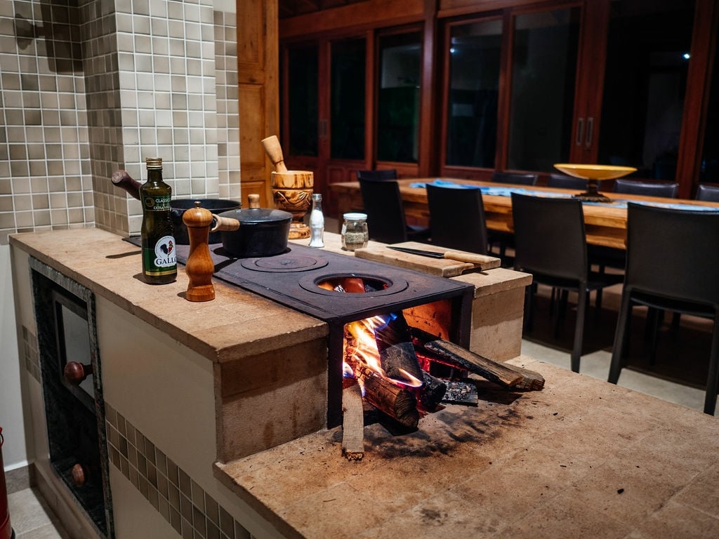 An outdoor cooking area is featured, showcasing a traditional wood-burning stove with a flame visible. Nearby, a wooden table is set for dining, surrounded by black chairs. Natural light enters through large windows, creating a warm and inviting space for gathering.