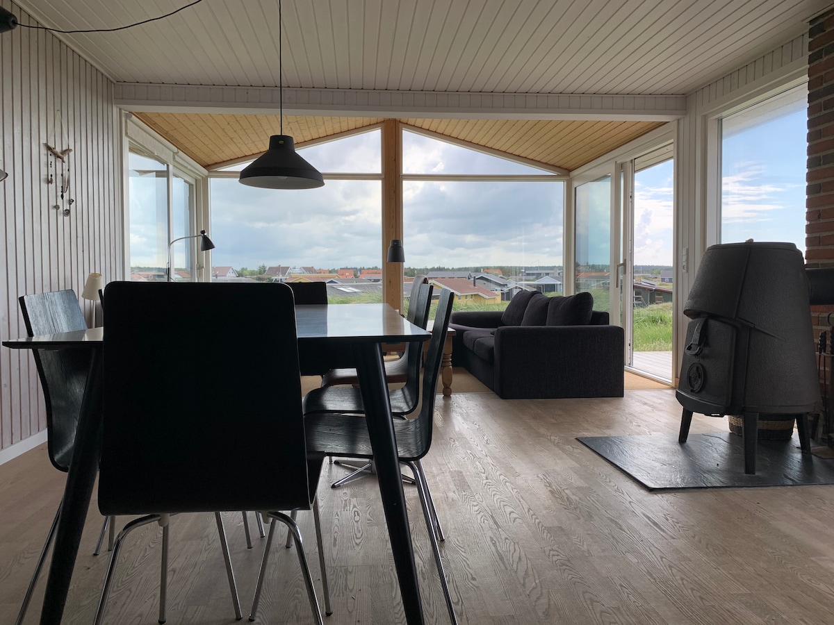 A spacious dining area is visible, featuring a black dining table surrounded by sleek chairs. Large windows offer expansive views of the surrounding landscape, allowing natural light to fill the room. A cozy seating area is positioned nearby, complete with a dark sofa and a wood-burning stove.