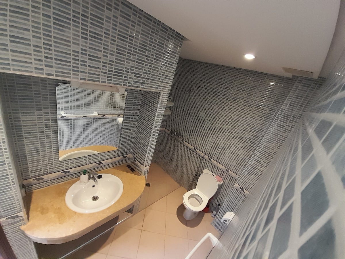 A modern bathroom is featured with a sleek vanity and sink, complemented by a large mirror. The walls are lined with patterned tiles, and a light-colored floor tile adds contrast. A toilet is positioned against one wall, providing a practical use of space.
