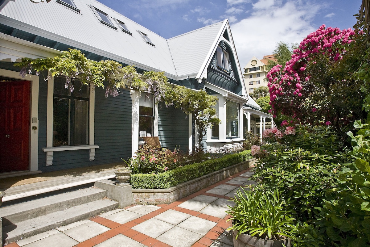 A charming heritage home is framed by vibrant gardens, featuring colorful flowers and lush greenery. A welcoming pathway of stone tiles leads to the entrance, highlighted by an inviting red door. The structure showcases elegant architectural details, including a steeply pitched roof and large windows.