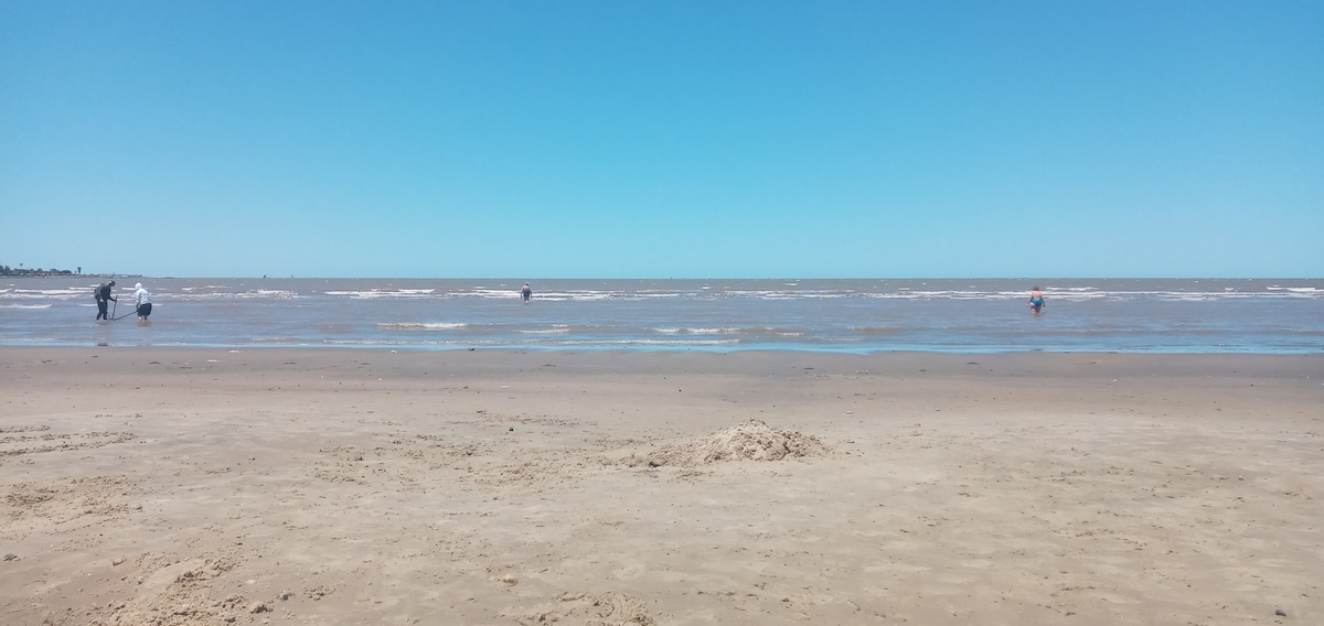 A sandy beach stretches under a clear blue sky, with gentle waves lapping at the shore. Figures can be seen wading into the water from the beach, while soft sand creates a smooth foreground undisturbed by footprints.