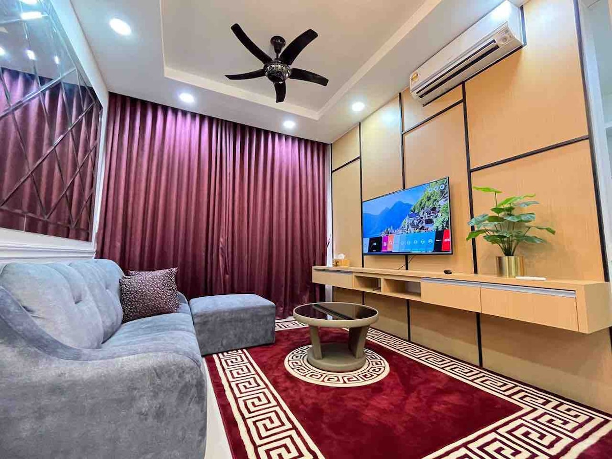 A comfortable living area features a gray sectional sofa complemented by decorative pillows. A low table sits in front of the sofa on a plush red area rug, while a wall-mounted TV displays vibrant visuals. Dark curtains frame the windows, and a potted plant adds a touch of greenery.
