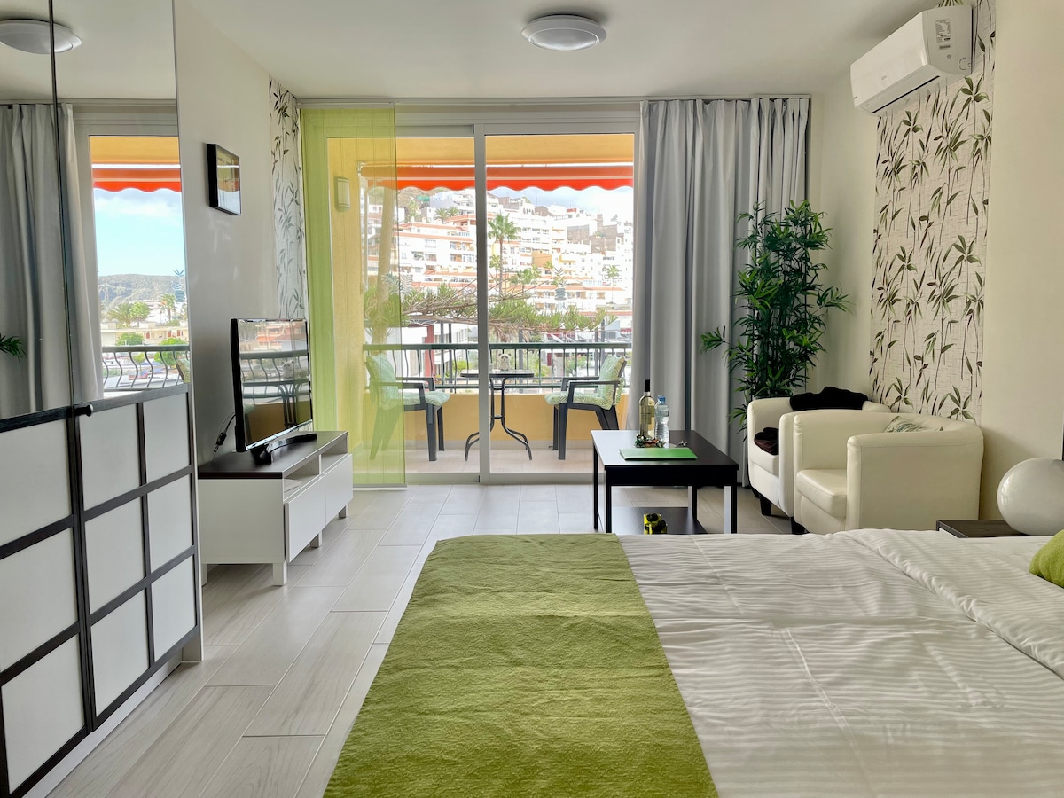A spacious studio is showcased, featuring a comfortable bed with a green accent blanket. A seating area includes a white armchair and a small table. Large glass doors lead to a balcony, providing views of the surrounding area and natural light.