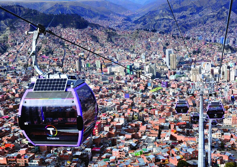 A series of gondolas traverse above a sprawling cityscape, showcasing a vibrant mix of buildings and rooftops. The towering mountains and expansive view of La Paz provide a dramatic backdrop, with clear skies allowing for a bright and expansive sight.