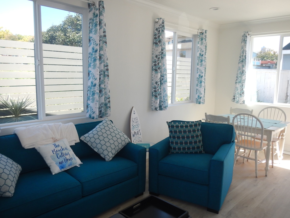 A bright and airy living area features a blue couch and a matching armchair, complemented by decorative pillows. Light streams through two large windows adorned with floral curtains. A dining table with white chairs is visible in the background, enhancing the inviting atmosphere.