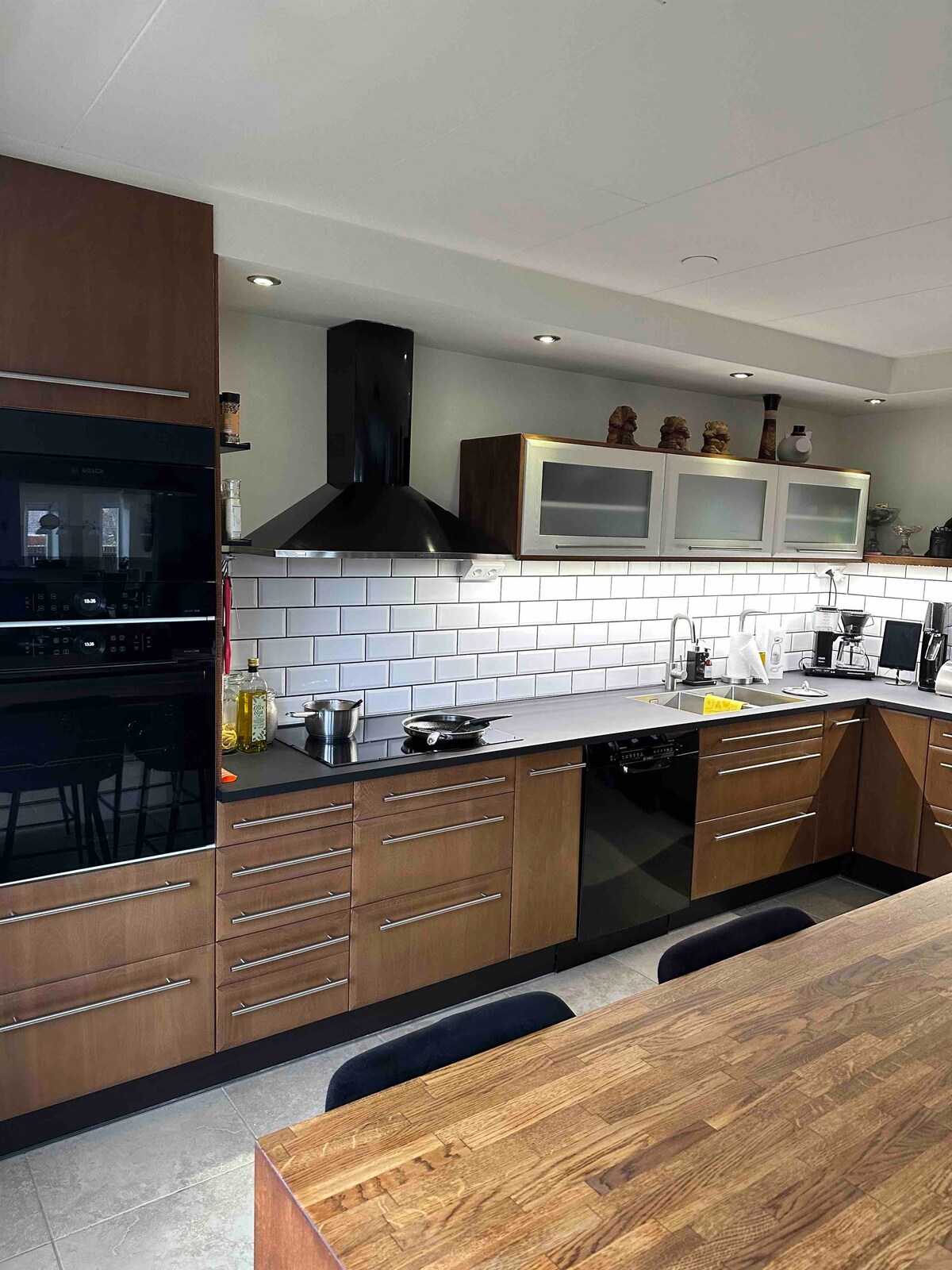 A modern kitchen is presented with sleek wooden cabinetry and a contrasting white tiled backsplash. Stainless steel appliances, including an oven and hood, are visible. A spacious wooden dining table is positioned nearby, complementing the kitchen's contemporary design.