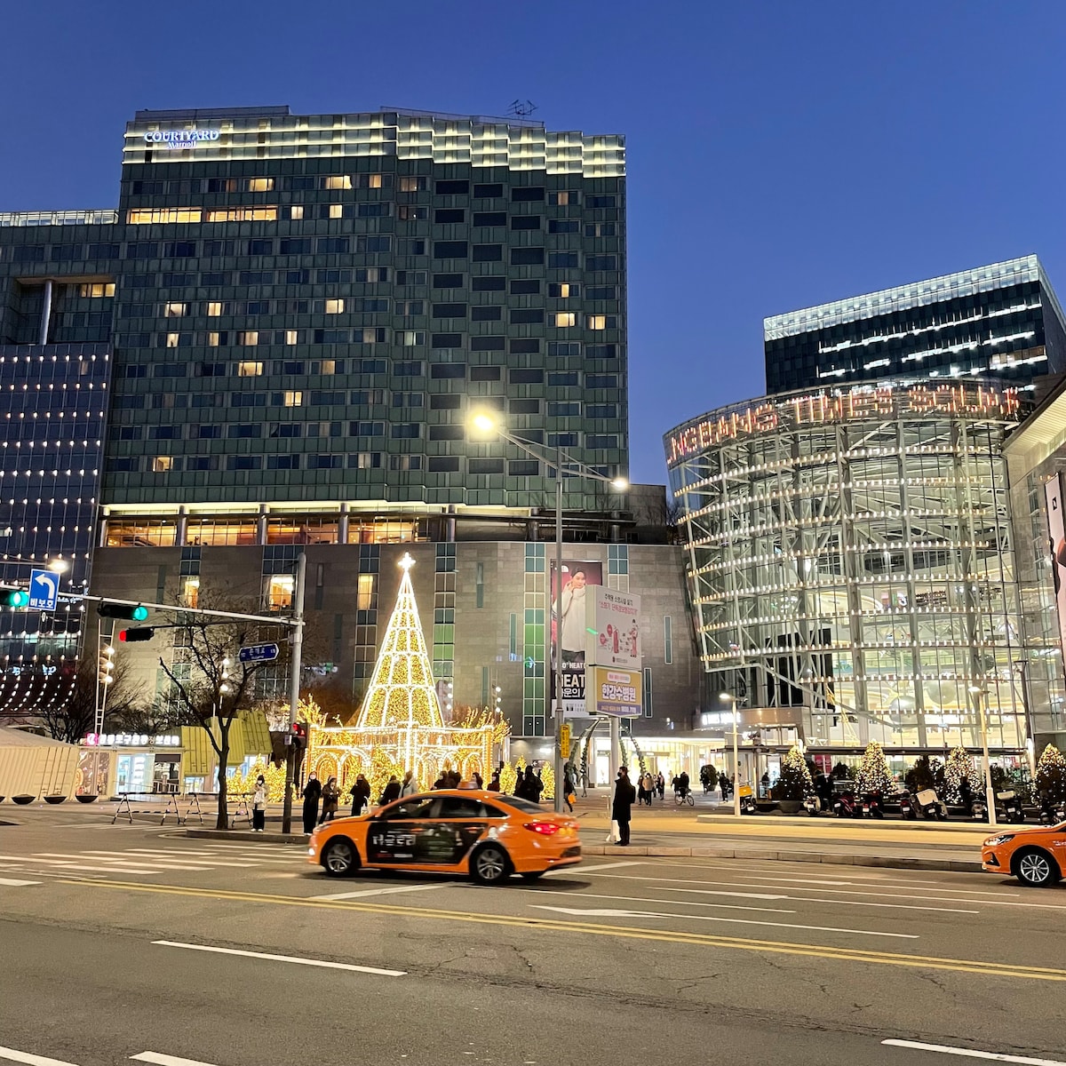 The image depicts a nighttime view of a vibrant cityscape featuring Times Square. A tree decorated with lights stands prominently at the forefront. Nearby, taxis are seen passing by, while modern buildings create an urban backdrop illuminated by lights.