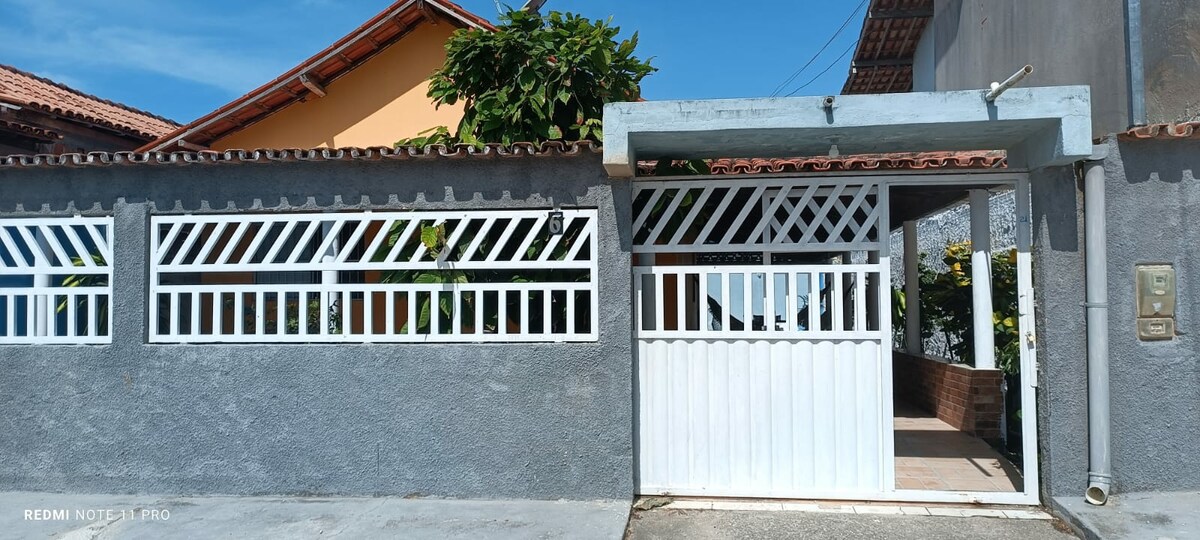 A gated entrance is presented with a white wooden door and a decorative grille. The walls are finished in a subtle gray tone, while lush green plants are visible through the grates, adding a touch of nature to the welcoming façade.