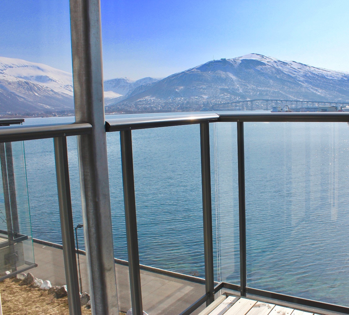 A glass-railed balcony offers views of a calm, glistening waterfront, framed by snow-capped mountains in the distance. The clear blue sky complements the serene landscape, inviting relaxation and connection with nature.