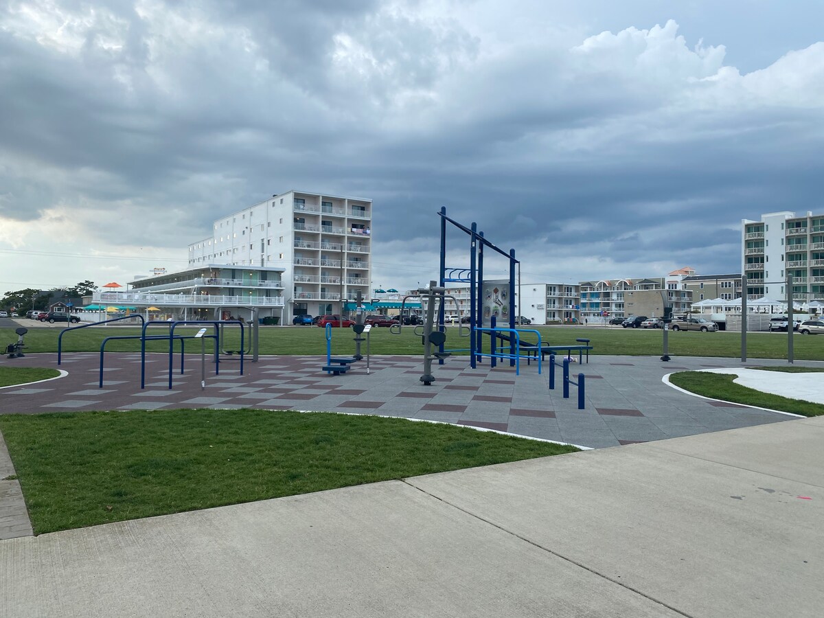An open grassy area features a modern play space equipped with fitness equipment and a soft surface for safety. In the background, several multi-story buildings line the street, indicating a vibrant community atmosphere. The sky displays a mix of clouds, hinting at a dynamic weather pattern.