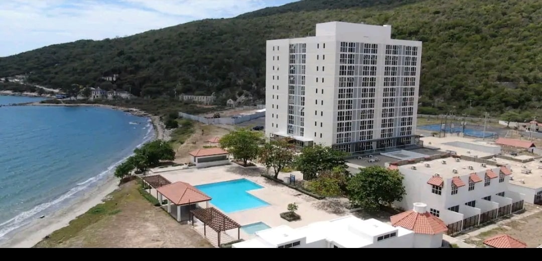 An aerial view presents a modern high-rise building adjacent to a serene coastline. A pool is situated in a landscaped area, complemented by minimalist outdoor structures. Lush greenery extends towards the mountains in the background, highlighting the tranquil environment.