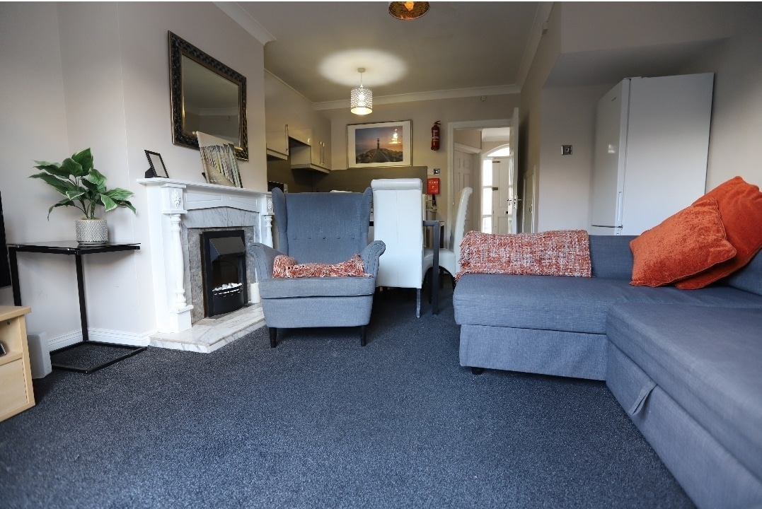 A welcoming living area features a gray sectional sofa and two armchairs with orange throw pillows. A white fireplace adds a central focus, while a small plant sits on a side table. The space is illuminated by natural light from nearby windows.