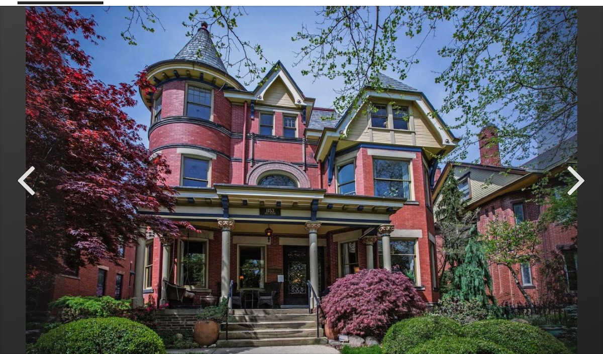 The exterior of a historic Victorian house is depicted, showcasing rich red brick and intricate architectural details. Lush greenery surrounds the entrance, with a wooden porch supported by decorative columns. Windows are adorned with colorful shutters, complementing the vibrant foliage in the foreground.