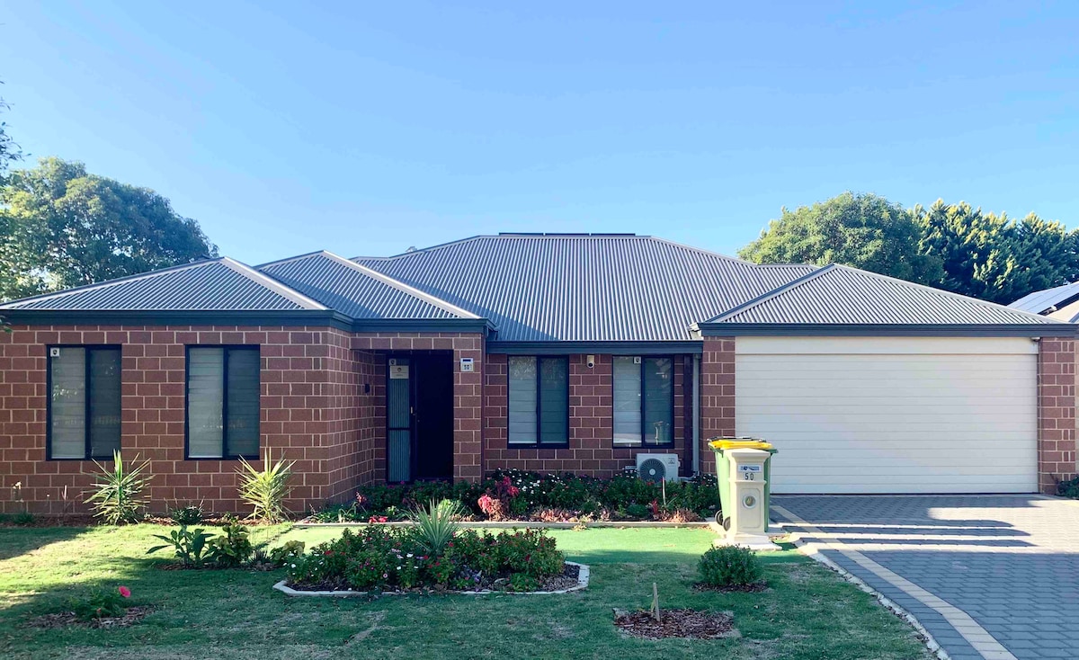 The exterior features a well-maintained brick facade with a dark roof. A manicured lawn with small shrubs and flowers is visible. The driveway leads to a spacious garage with a door. Large windows reflect natural light, and a path leads to the front entrance.