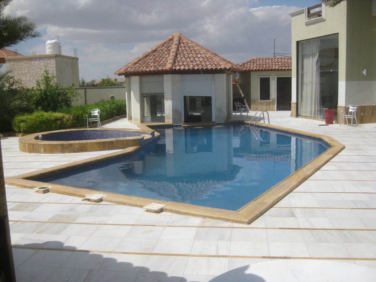 A swimming pool with a distinctive shape is surrounded by a spacious stone patio, featuring a separate spa area. Sun loungers are positioned nearby, and a covered seating area with windows provides a shaded retreat. Lush greenery is visible in the background, complementing the peaceful setting.