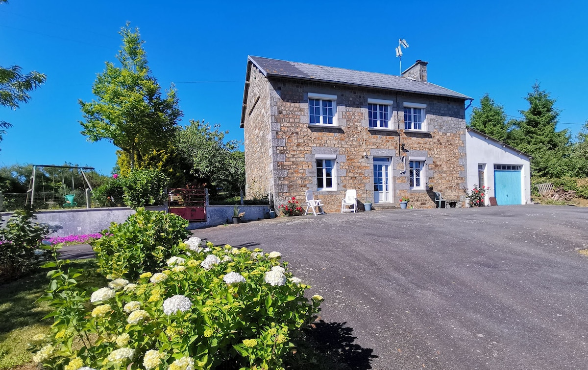 A well-maintained stone house is positioned in a quiet area, surrounded by greenery and flowering plants. The exterior features a large driveway leading up to the entrance, with seating options visible nearby. A garage is attached to the house, contributing to the overall spaciousness.