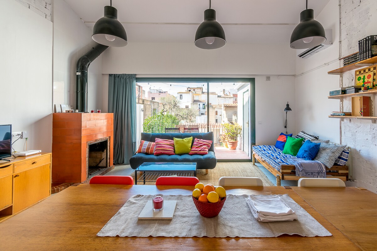 A bright living space is showcased, featuring large glass doors that open to a terrace. A dining table with a light tablecloth is placed in the foreground, surrounded by colorful chairs. A cozy seating area includes a sofa and a daybed, with a variety of cushions.