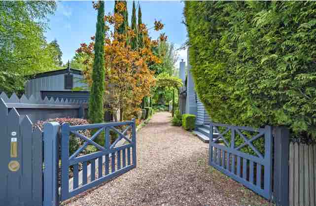 A well-maintained pathway is visible through an open gate, framed by lush greenery and flowering trees. The gravel path leads towards the house, flanked by tall hedges that provide a sense of privacy and seclusion.