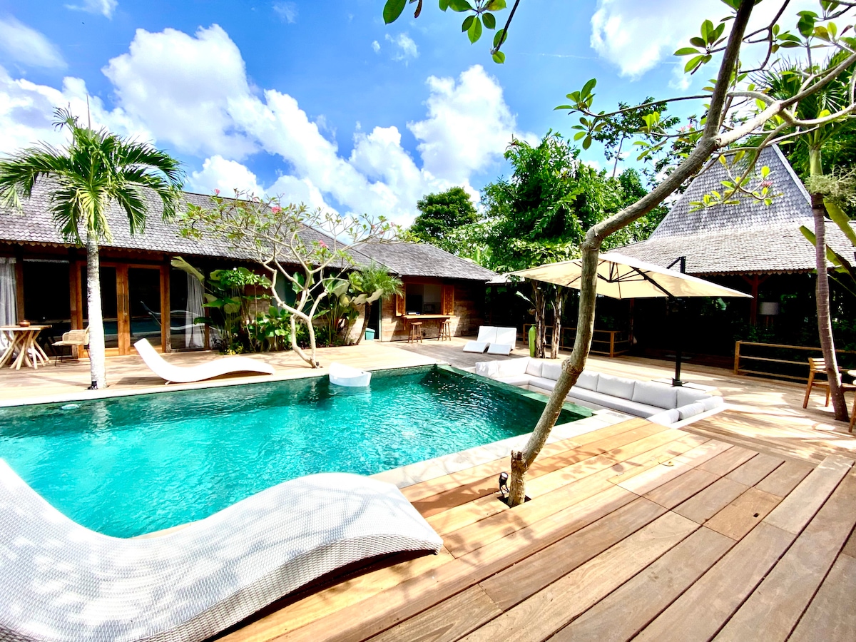 A large swimming pool is surrounded by wooden decking and tropical greenery. Lounge chairs are positioned around the pool, offering a place to relax. Sun umbrellas provide shade near the deck. The sky above is bright with scattered clouds.