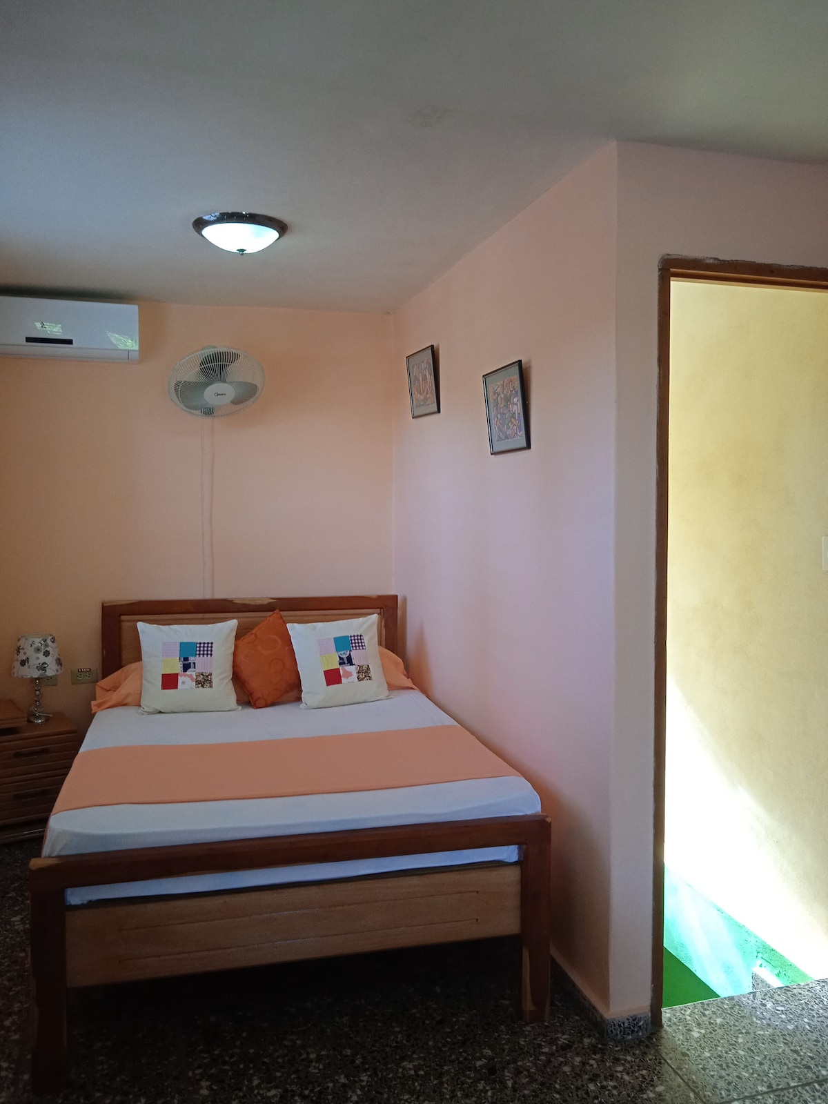 The room features a well-lit sleeping area with soft peach walls and a spacious bed adorned with decorative pillows. A bedside table and air conditioning unit are visible, while a door leads to another space, adding convenience to the layout.