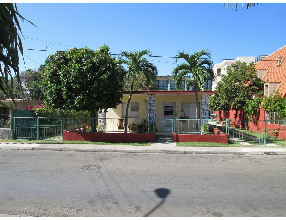 The exterior of a residential house is displayed, featuring a front yard with green grass and palm trees. A fence surrounds the property, and a pathway leads to the entrance. Nearby, other houses and foliage are visible, creating a serene neighborhood atmosphere.