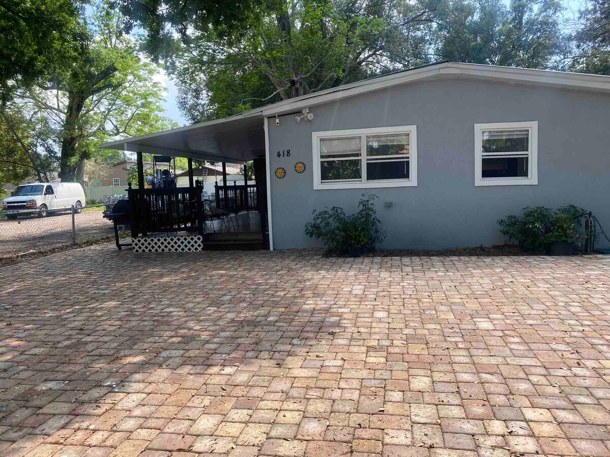 A single-story house with a light blue exterior is surrounded by green trees. The front features a spacious, paved area with a stone pattern and a covered porch, which includes multiple seating options, offering a shaded spot for relaxation.