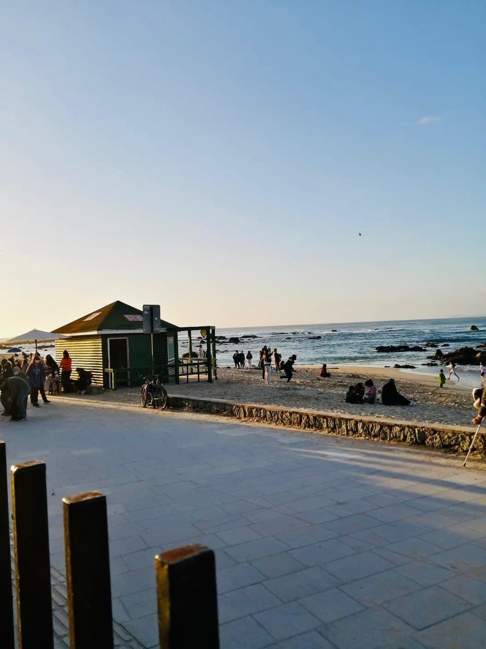 A beach scene captures visitors enjoying the shoreline during sunset. A small green building is visible, and people are scattered along the sandy beach. Waves gently lap against the shore, with a clear sky adding to the tranquil atmosphere.