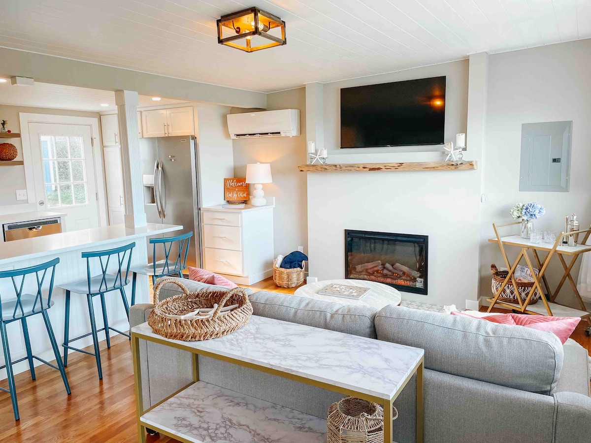 The living area is equipped with a 50-inch mounted smart TV and a cozy electric fireplace beneath a wooden mantle. A comfortable sectional sofa is positioned around a modern coffee table, with a breakfast bar featuring three stools visible in the background. Natural light fills the space.