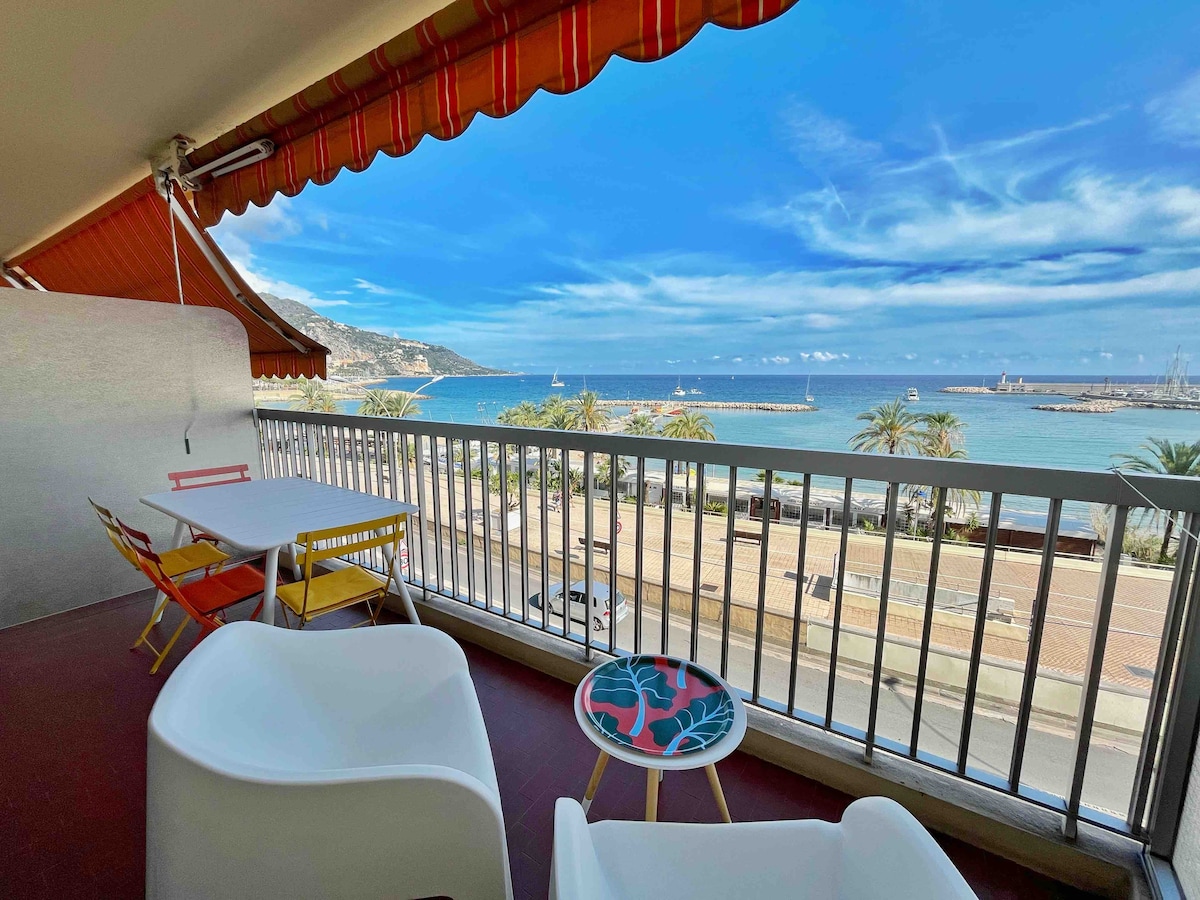 A balcony offers a view of the Mediterranean Sea and the bustling beach below. Two chairs and a small table are positioned on the balcony, bordered by decorative metal railings. The sky features light clouds, and palm trees are visible in the foreground.