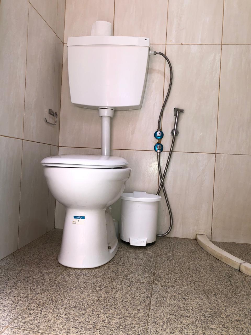 A clean and simple bathroom corner is shown, featuring a white toilet and a standard flush tank. A small trash bin sits nearby, while a shower spray attachment is positioned on the wall. The floor is tiled with light-colored stone, enhancing the space's functionality.