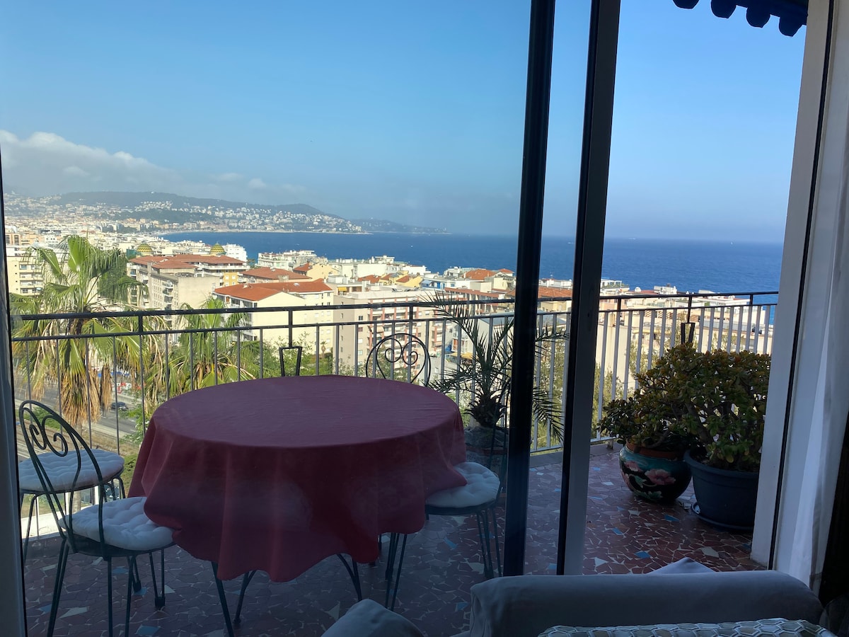 An outdoor balcony offers a view of the sea and distant hills, framed by sliding glass doors. A round table with four chairs is positioned on the balcony, complemented by potted plants and palm trees. The scene captures a serene coastal atmosphere.