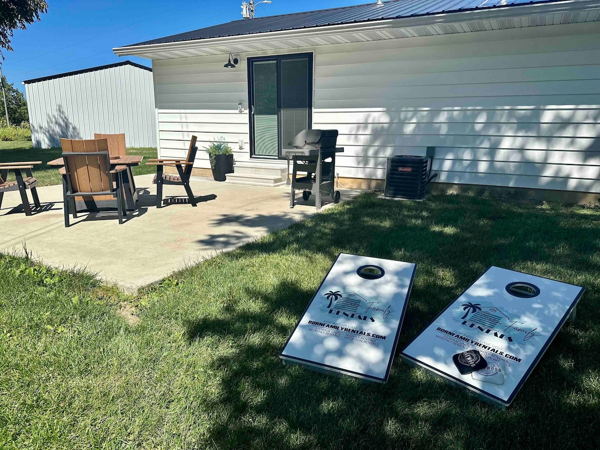 An outdoor patio area includes a dining set and a grill, surrounded by lush green grass. Two cornhole boards are positioned in front of a cottage, inviting outdoor games. A door leads to the interior, and a large shed is visible in the background.