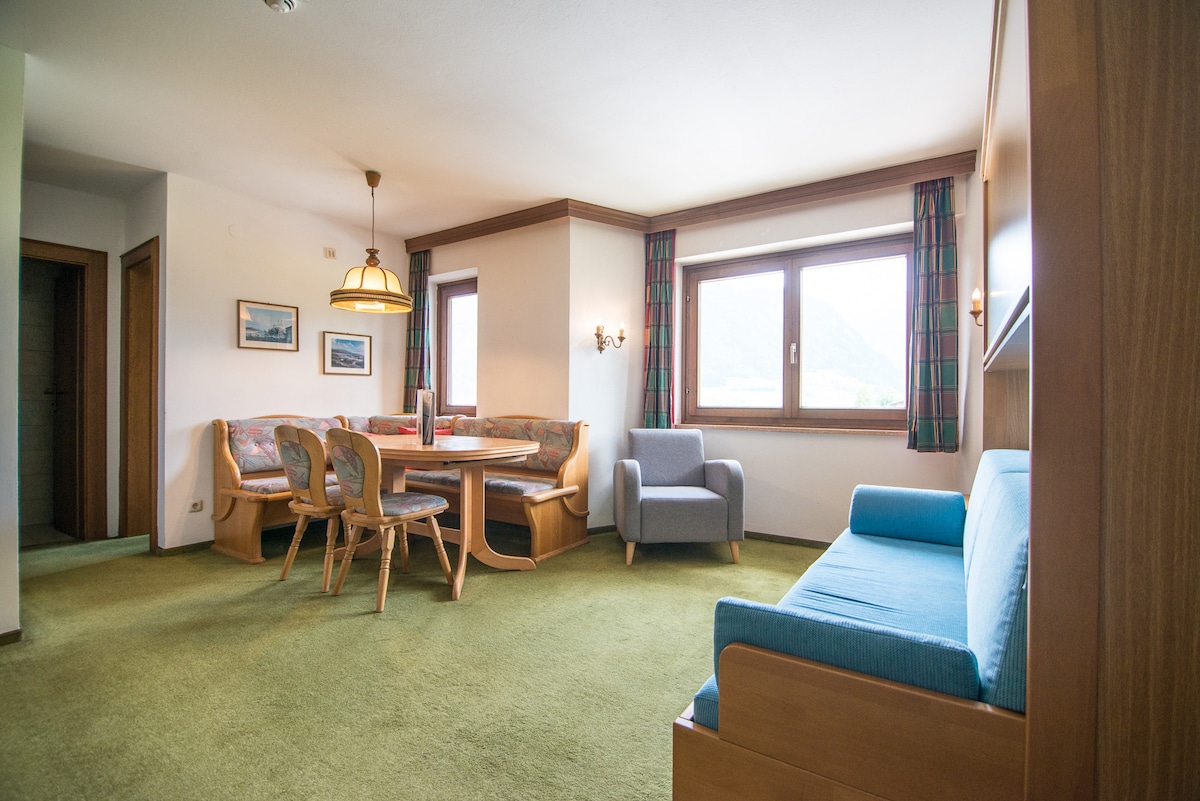 A spacious living area features a comfortable corner sofa and a light blue armchair. A dining table with four wooden chairs is positioned next to large windows, offering views of the outdoor landscape. The room is accented with subtle green carpeting and light wood paneling.