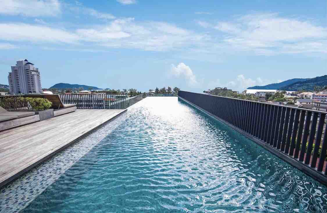 A double infinity pool extends towards the horizon, framed by wooden decking and a railing. The clear water reflects the sky, creating a serene atmosphere. Nearby, loungers can be seen, providing spaces for relaxation while enjoying sweeping views of the surrounding landscape.
